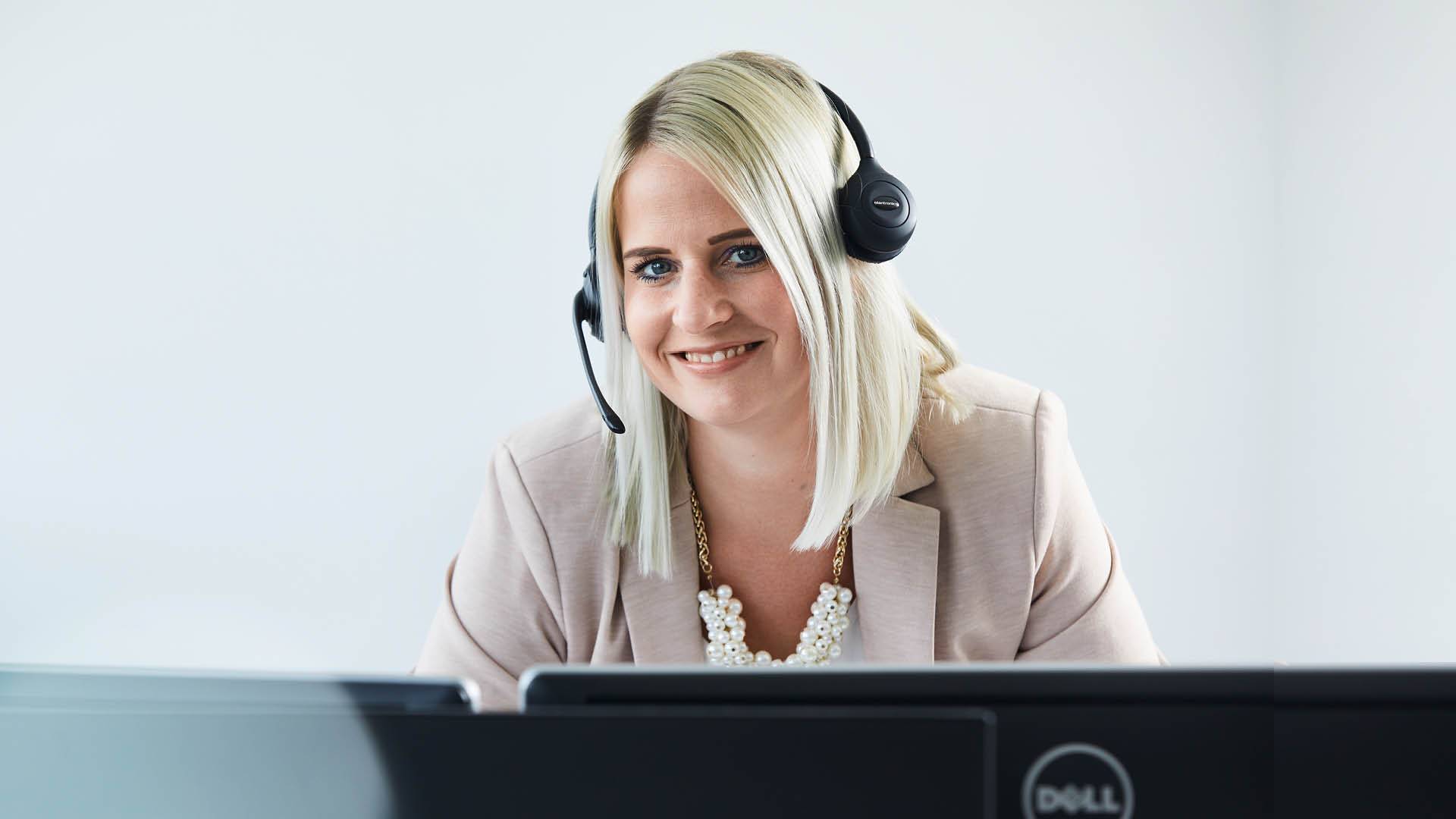 Eine Mitarbeiterin mit Headset aus dem Bereich IT lächelt von ihrem Arbeitsdplatz.